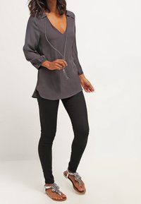 Graue, fließende Bluse mit tiefem V-Ausschnitt und dreiviertel Ärmeln, kombiniert mit schwarzen Skinny-Jeans und verzierten weißen Sandalen.