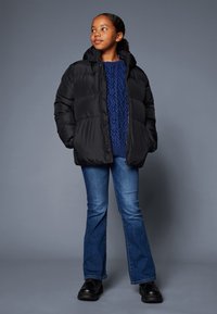 Doudoune noire avec capuche, présentant un design matelassé. Portée par-dessus un pull en coton bleu à mailles et un jean bleu évasé, associée à des chaussures noires.