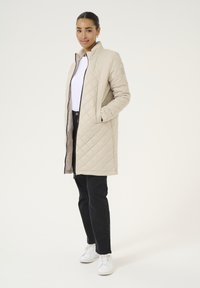 Beige gesteppter Mantel mit hohem Kragen, vorne Reißverschluss und zwei Seiten Taschen, kombiniert mit schwarzen Hosen und weißen Sneakers. Schlichtes Design.