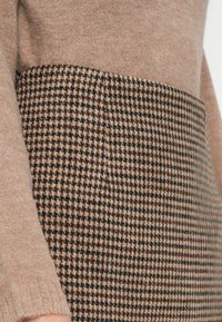 Pletený svetr s lehkým béžovým vrškem a spodní částí wovenu s houndstooth vzorem v černé a hnědé barvě. Měkká textura, uvolněný střih.