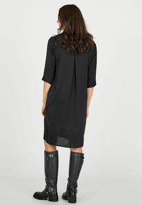 Vestido negro de corte recto con mangas tres cuartos, que presenta un detalle de pliegue en la parte posterior. Combinado con botas de cuero negro hasta la rodilla. Textura suave.