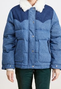 Veste rembourrée bleue avec un empiècement bleu marine, col en polaire blanche, boutons-pression, deux poches avant, portée sur un pantalon vert foncé.
