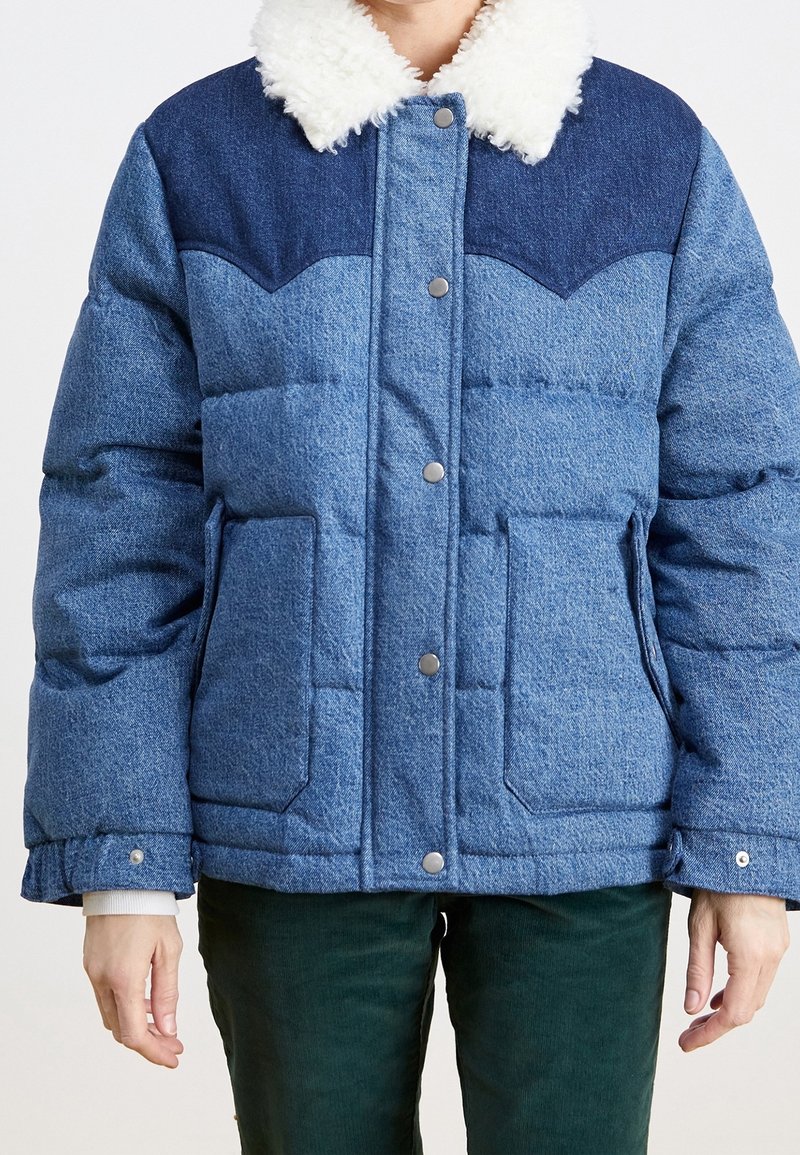 Veste rembourrée bleue avec un empiècement bleu marine, col en polaire blanche, boutons-pression, deux poches avant, portée sur un pantalon vert foncé.
