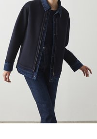 Marinblå zip-up jacka med kontrasterande denimkrage och manschetter, gjord av mjukt tyg. I kombination med mörkblå jeans för en enhetlig look.