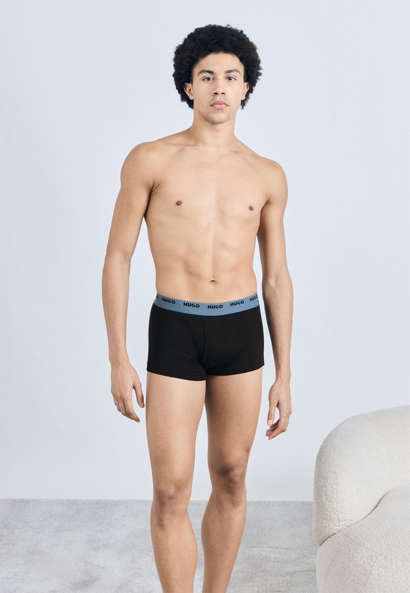 Boxers de algodão pretos com um cós azul com o logótipo "HUGO", apresentando um ajuste justo e design minimalista. Textura suave sem padrões.