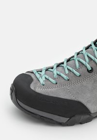Chaussure de randonnée en nubuck gris avec des lacets turquoise, un capuchon en caoutchouc à l'avant et une semelle noire texturée pour une meilleure adhérence. Nom de la marque "SCARPA" embossé sur le côté.