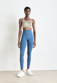 Kvinne som står i beige sports-bh, høytlivde blå leggings og hvite joggesko mot en enkel hvit vegg.