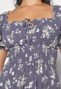 Kvinde iført lilla kjole med hvidt blomsterprint, puffede ærmer, firkantet udskæring og indsnævret talje med bundet sløjfe.