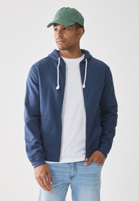 Mann som har på seg grønn caps, marineblå hettegenser over hvit t-skjorte, lyseblå jeans, står med en hånd i lommen og ser til høyre.