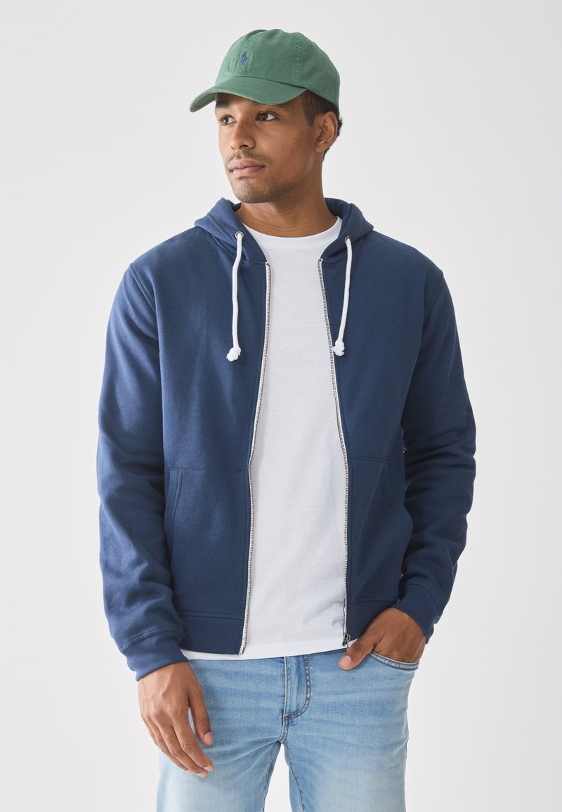 Mann som har på seg grønn caps, marineblå hettegenser over hvit t-skjorte, lyseblå jeans, står med en hånd i lommen og ser til høyre.