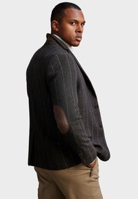 Blazer in lana grigia scura con righe verticali, toppe in pelle marrone sui gomiti e vestibilità sartoriale. Caratterizzato da chiusura a due bottoni e rever a punta.