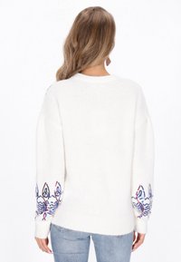 Fehér kötött pulóver hosszú ujjakkal, amelyen bonyolult kék és piros hímzett minták találhatók az ujjak végén. Puha tapintású és laza szabású.