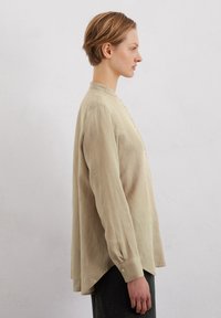 Beige linen shirt with a round collar, vertical button closure, and long sleeves. Features a relaxed fit and rounded hem at the back.