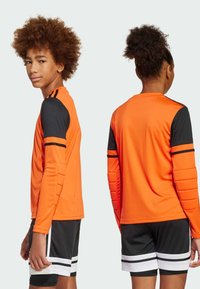 Oranje sporttrui met lange mouwen en zwarte mouwen, versierd met gewatteerde accenten. Gecombineerd met zwarte shorts met witte en zwarte strepen.