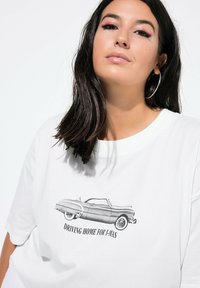 Witte katoenen T-shirt met een vintage auto-illustratie in het zwart en de tekst "RIJDEN NAAR HUIS VOOR KERST." Casual fit en ronde halsontwerp.