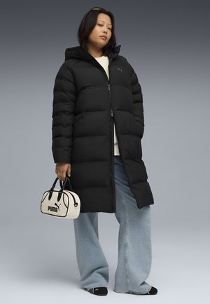 Zwarte pufferjas met een capuchon, geborduurd ontwerp en een rits aan de voorkant. Gecombineerd met losse blauwe jeans en een witte handtas met zwarte accenten.