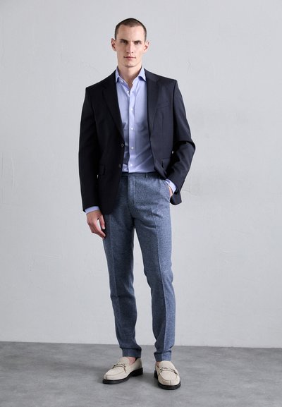 Blazer bleumarin peste o cămașă lila deschis, asociat cu pantaloni gri texturat și loafers bej cu detalii din lanț. Design simplu, croială, aspect formal.