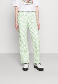 Calças largas verde e brancas com riscas, apresentando um padrão ondulado, feitas de tecido leve, combinadas com sapatos brancos e uma t-shirt gráfica.