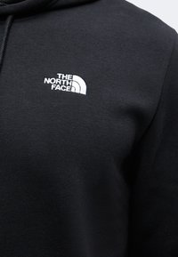 Sweat à capuche noir avec logo The North Face brodé en blanc sur la poitrine, montrant la texture du tissu et une partie du cordon de la capuche.