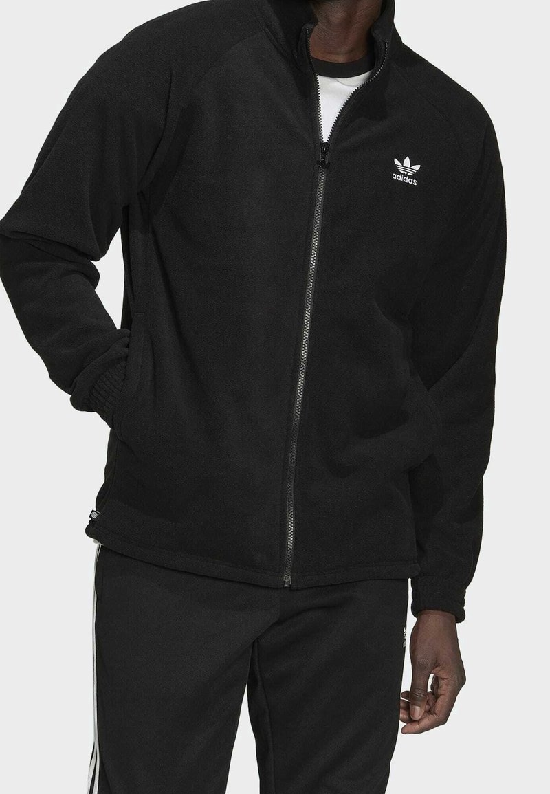 Svart fleecejacka med dragkedja fram, ribbade muddar och två sidofickor. Har Adidas-logotyp i vitt på vänstra bröstområdet.
