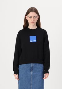 Felpa nera con una grafica a forma di quadrato blu e logo sul petto. Presenta polsini e orlo a coste. Indossata con una gonna di jeans.