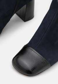 Bottine en daim bleu marine avec un bout en cuir verni noir brillant, un talon carré et une fermeture éclair latérale.