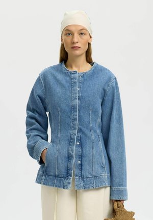 Femme au visage sérieux portant une veste en jean boutonnée, un pantalon crème, un foulard blanc sur la tête, tenant un sac tissé en forme d'étoile.