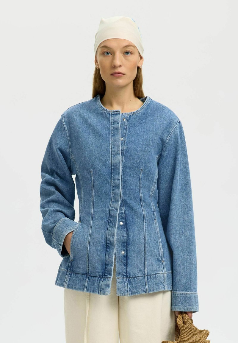 Femme au visage sérieux portant une veste en jean boutonnée, un pantalon crème, un foulard blanc sur la tête, tenant un sac tissé en forme d'étoile.