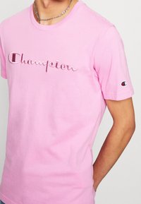 Růžové bavlněné tričko s vyšitým logem "Champion" v bílé a červené barvě. Krátké rukávy s nášivkou loga na levém rukávu.