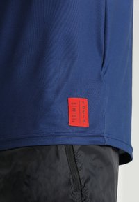 Blue fabric with a small red rectangular label showing "TOKYO" and Japanese characters, paired with black textured pants with a zipper pocket.