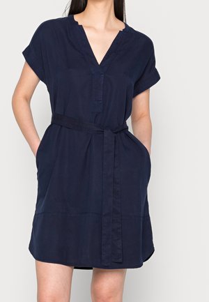 Frau trägt ein kurzärmeliges marineblaues Kleid mit einem in der Taille gebundenen Gürtel und hat die Hände in den Taschen vor einem einfarbigen hellen Hintergrund.