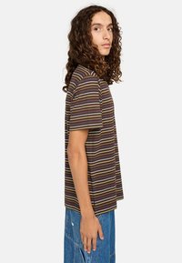 T-shirt rayé avec une base marron et des rayures horizontales jaunes, bleues et blanches. Manches courtes et coupe décontractée. Porté avec un jean en denim bleu.