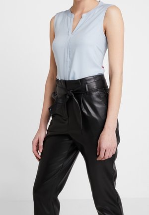 Mujer con blusa sin mangas azul claro metida en pantalones de cuero negros de cintura alta con cinturón anudado, de pie contra un fondo blanco.