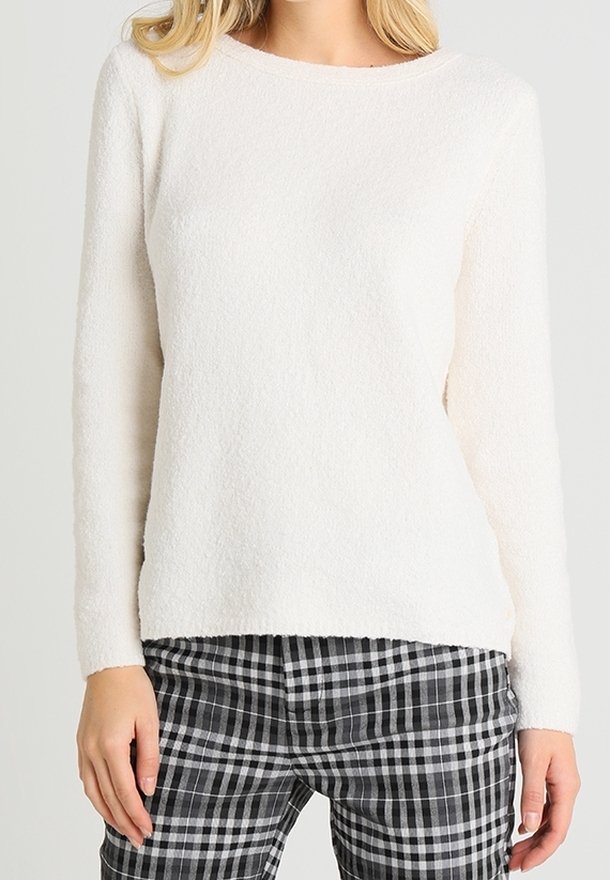 Personne portant un pull blanc à manches longues et à texture douce ainsi qu’un pantalon à carreaux noir et blanc, debout devant un fond uni.