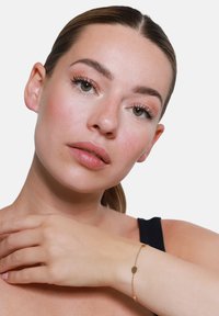 Bracciale in tonalità oro con piccoli anelli ovali e rotondi, elegantemente fissato su un polso con una pelle liscia e tonalità abbronzata.