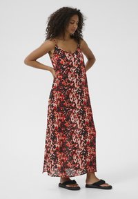 Robe maxi en tissu léger avec un motif abstrait rouge et noir, dotée de fines bretelles et d'une coupe décontractée. Portée avec des sandales noires.