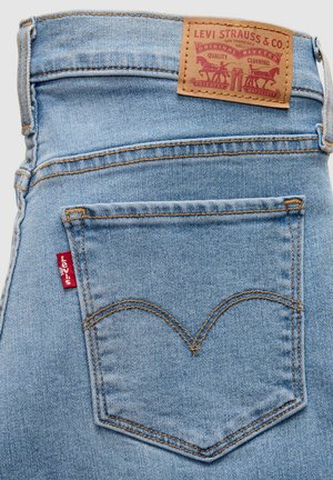 Lyseblå denimjeans med en mærket læderlap, en rød mærkat på baglommen og karakteristisk syning i et buet mønster.