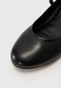 Gros plan sur la partie avant d'une chaussure ballerine en cuir noir avec un bout arrondi et une texture lisse sur un fond clair.