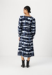 Maxikjole i marineblå med hvit tie-dye mønster. Har lange ermer, samlet bak og en flytende silhuett. Brukes med svarte ankelstøvletter.