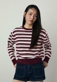 Sweter w paski w kolorze burgundowym i kremowym, z ribbowanym dekoltem crew, długimi rękawami, zestawiony z ciemnoniebieskimi szortami dżinsowymi. Gładki materiał, casualowy styl.