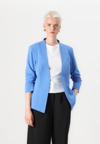 Svijetloplavi sako krojenog stiliziranja s dugim rukavima, strukturiranim dizajnom i otvorenim prednjim dijelom. Nosivo preko bijele bluze i crnih širokih hlača.