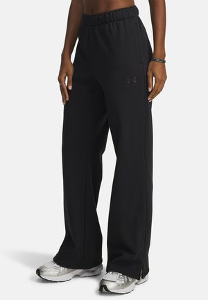 Person iført løse sorte Under Armour bukser med elastisk talje og sølvfarvede sportssko, stående mod en ensfarvet baggrund.