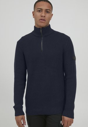 Navyblauer Strickpullover mit hohem Kragen, halbem Reißverschluss, gerippten Bündchen und Saum. Enthält ein strukturiertes Muster und ein Logopatch am Ärmel.
