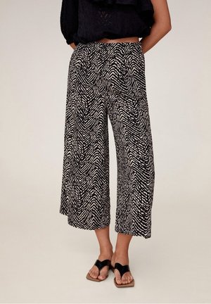 Kobieta ubrana w czarne i beżowe szerokie spodnie o kroju culottes w azteckie wzory, połączone z czarnymi sandałami na płaskim obcasie z pętelką na palce, stojąca na tle jednolitej ściany.