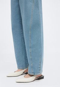 Άτομο που φοράει ανοιχτό μπλε τζιν με ευθείες γραμμές και λευκά slingback παπούτσια με μυτερό μπροστινό μέρος και χαμηλά μαύρα τακούνια σε λευκό φόντο.