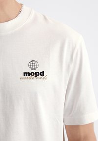 Biela bavlnená tričko s okrúhlym výstrihom, krátkymi rukávmi a prednou grafikou globusu a textom "mopd world.wide" v čiernej a zlatej farbe.