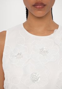 Gros plan d'une personne portant un haut blanc avec de grandes fleurs brodées et des détails en sequins au niveau de la poitrine.