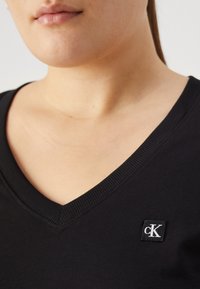 Černé bavlněné tričko s výstřihem do V. Obsahuje malou čtvercovou nášivku s logem "CK" v bílé barvě na levé části hrudi.