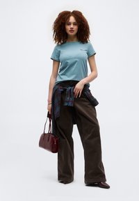 Νεαρή γυναίκα με σγουρά μαλλιά που φορά μπλε μπλούζα, σκούρες ευρείες παντελόνες, καρό πουκάμισο δεμένο στη μέση, κρατά μπορντό τσάντα.