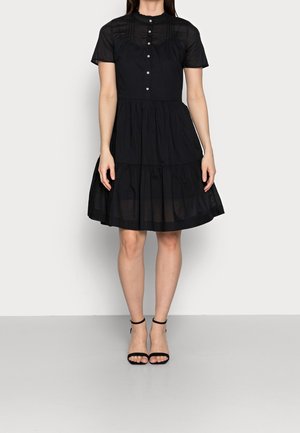Mujer con vestido corto negro abotonado, mangas cortas y falda de volantes, combinado con sandalias negras de tacón alto con tira en el tobillo.